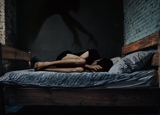 Imagem captura a essência arrepiante da paralisia do sono: uma pessoa encolhida de medo, com uma mão sombria pairando sobre a cabeça, simbolizando o desamparo e o terror da experiência. Metrópoles