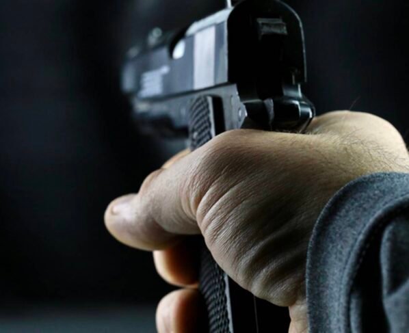 Imagem colorida de uma mão de uma pessoa branca segurando uma pistola