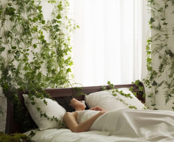 Mulher deitada em cama com jogo de cama branco e paredes do quarto cheias de plantas verdes