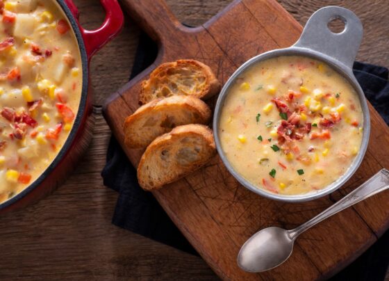 Imagem mostra uma tigela de sopa amarela e pedaços de pão em cima de uma tábua de madeira.
