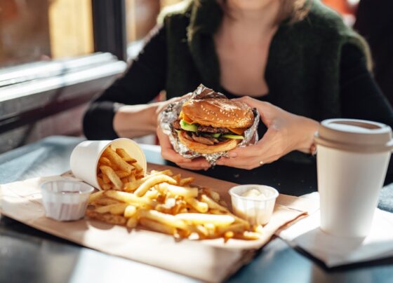 Close-up das mãos de uma mulher segurando um hambúrguer enquanto come batata frita. Metrópoles - calorias