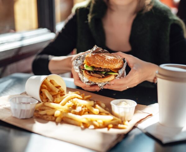Close-up das mãos de uma mulher segurando um hambúrguer enquanto come batata frita. Metrópoles - calorias