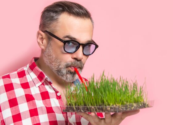 Foto colorida com fundo rosa, homem mais velho à frente bebendo água por um canudo diretamente da terra - Metrópoles.