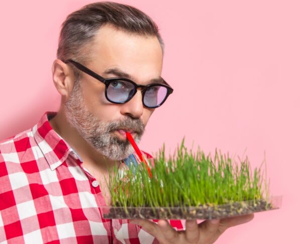 Foto colorida com fundo rosa, homem mais velho à frente bebendo água por um canudo diretamente da terra - Metrópoles.