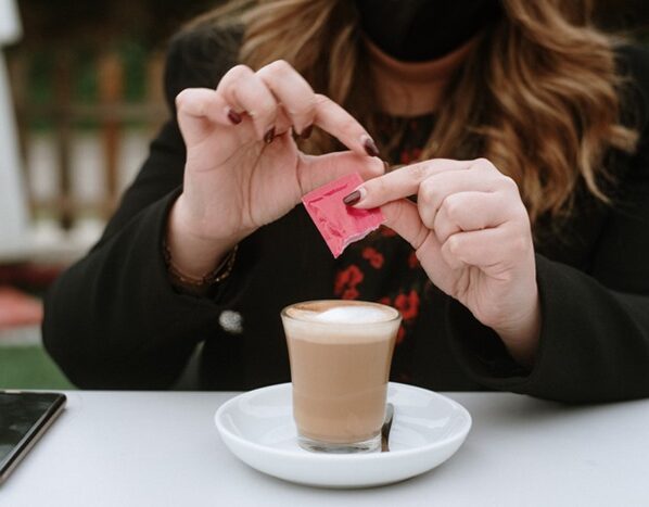 Imagem de mulher colocando adoçante em copo com bebida marrom - Metrópoles