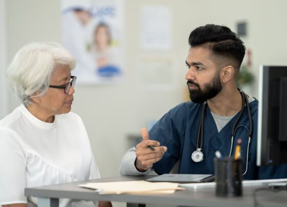 Foto colorida de um homem vestido de médico e uma senhora sendo atendida por ele em frente a um computdor - Metrópoles.