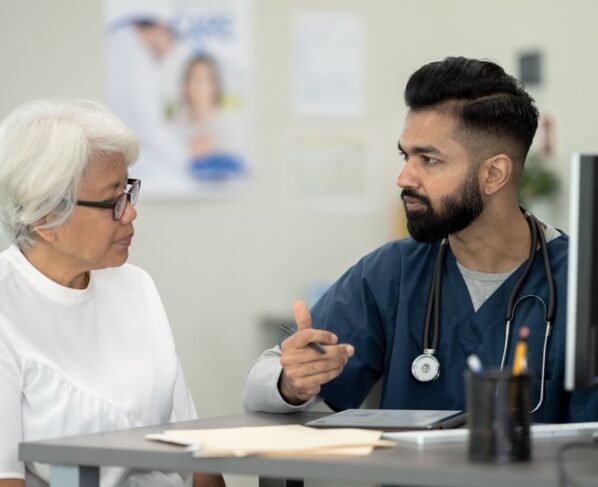 Foto colorida de um homem vestido de médico e uma senhora sendo atendida por ele em frente a um computdor - Metrópoles.