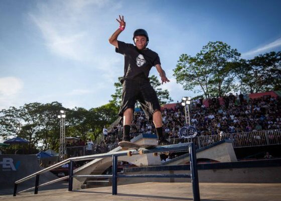 Um skatista em movimento salta com o skate sobre um obstáculo, capturado no ar sob luz intensa, com expressão concentrada e o público ao fundo.