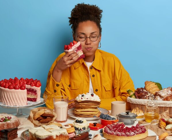 Foto colorida de mulher sentada atrás de uma mesa com muitas comidas, segurando um pedaço enorme de bolo com as mãos -Metrópoles.