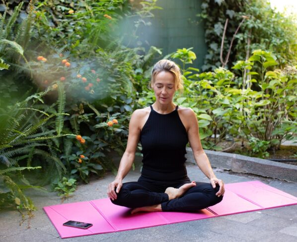 Imagem colorida de pessoa meditando em cima de tapete rosa em lugar verde - Metrópoles