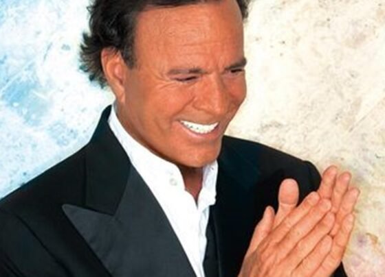 Julio Iglesias posa, sorridente, com as mãos juntas e blazer preto - Metrópoles