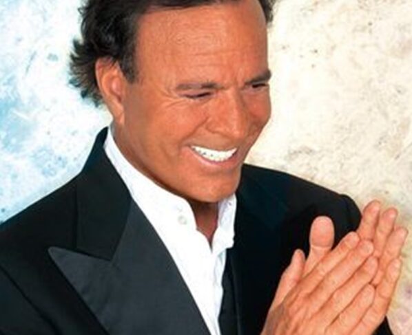Julio Iglesias posa, sorridente, com as mãos juntas e blazer preto - Metrópoles