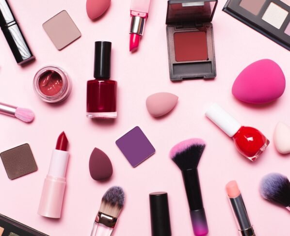 Conjunto de maquiagem com vista superior sobre fundo rosa pastel. Pincéis, batons, pó facial, blushes, esponjas, sombras e esmaltes. Metrópoles