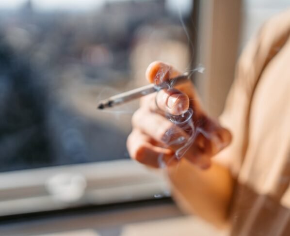 Close de um jovem/adolescente em pé perto da janela fumando um cigarro em casa. Metrópoles