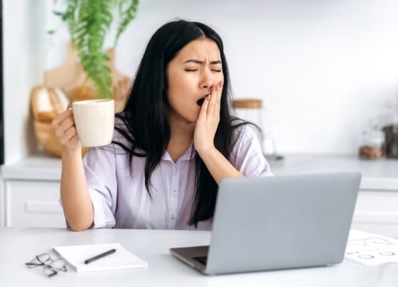 Garota asiática bonita e sonolenta, trabalhando remotamente, gerente de conteúdo, freelancer, estudante, sentada à mesa da cozinha com o laptop, começando o dia de trabalho, tomando café, bocejando, sem conseguir acordar. Metrópoles