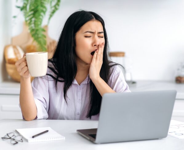 Garota asiática bonita e sonolenta, trabalhando remotamente, gerente de conteúdo, freelancer, estudante, sentada à mesa da cozinha com o laptop, começando o dia de trabalho, tomando café, bocejando, sem conseguir acordar. Metrópoles