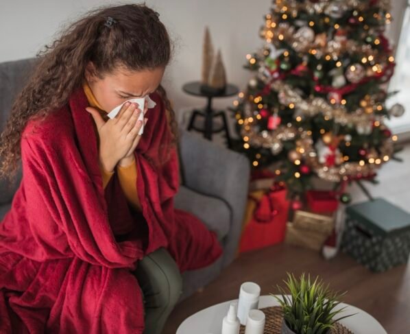 Jovem mulher sentada perto de uma árvore de Natal, coberta com um cobertor, assoando o nariz. Imunidade baixa no fim de ano. Metrópoles