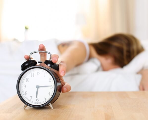 Imagem colorida mostra mulher loira deitada em uma cama desligando despertador - Metrópoles