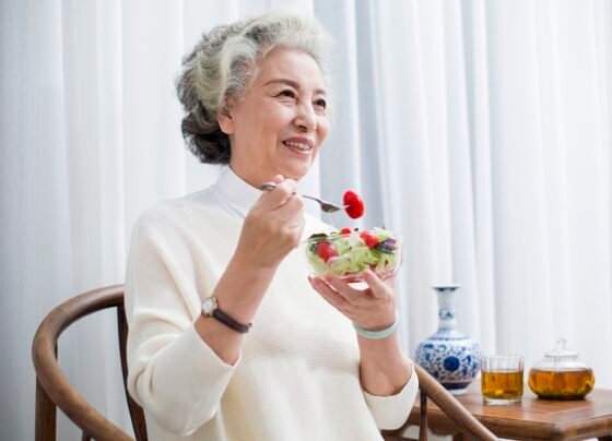 Foto colorida de mulher segurando tigela com frutas. Ela está levando garfo com fruta à boca - Metrópoles