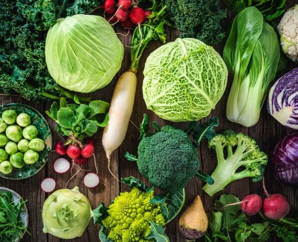 Variedade de vegetais crucíferos, incluindo repolho, brócolis, couve, nabo, couve-galega, romanesco, rabanete, rúcula, couve-rábano e rutabaga, disposta em tábua rústica de madeira. Metrópoles