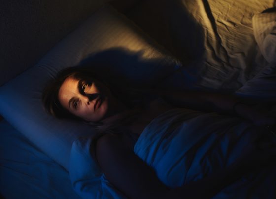 Foto colorida com fundo azul escuro e luz laranja, de mulher deitada na cama com olhos abertos, sintomas de insônia. - Metrópoles.
