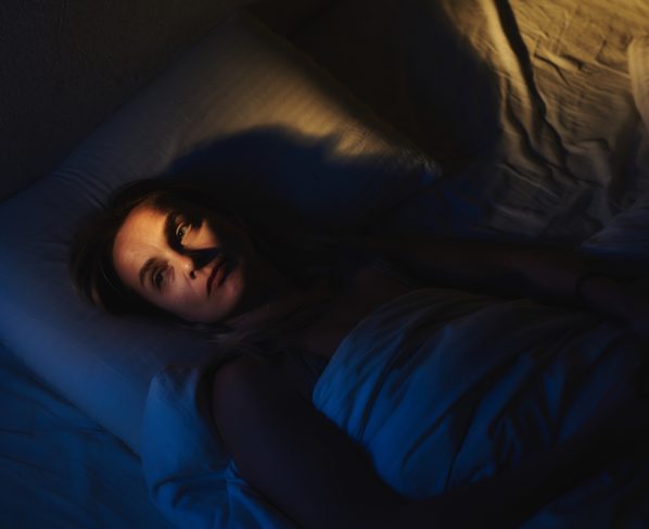 Foto colorida com fundo azul escuro e luz laranja, de mulher deitada na cama com olhos abertos, sintomas de insônia. - Metrópoles.